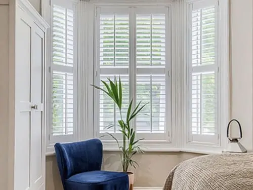 Bedroom shutters in a bay window with a plant and a blue chair.