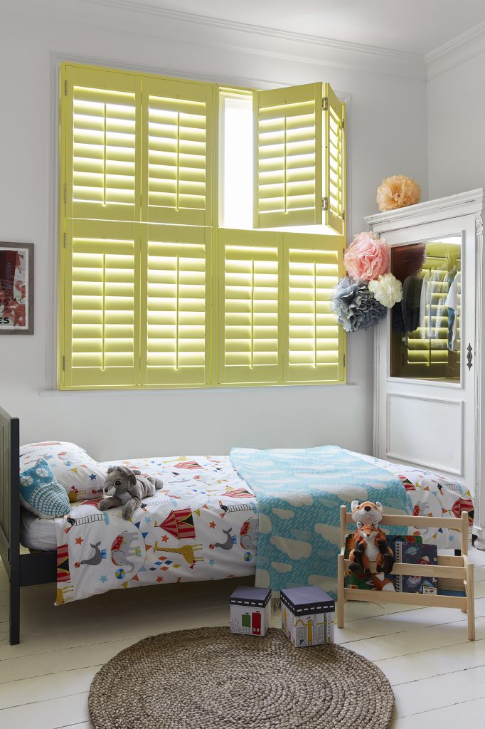 Yellow tier-on-tier window shutters in a children's room with a bed with circus patterns and toys.