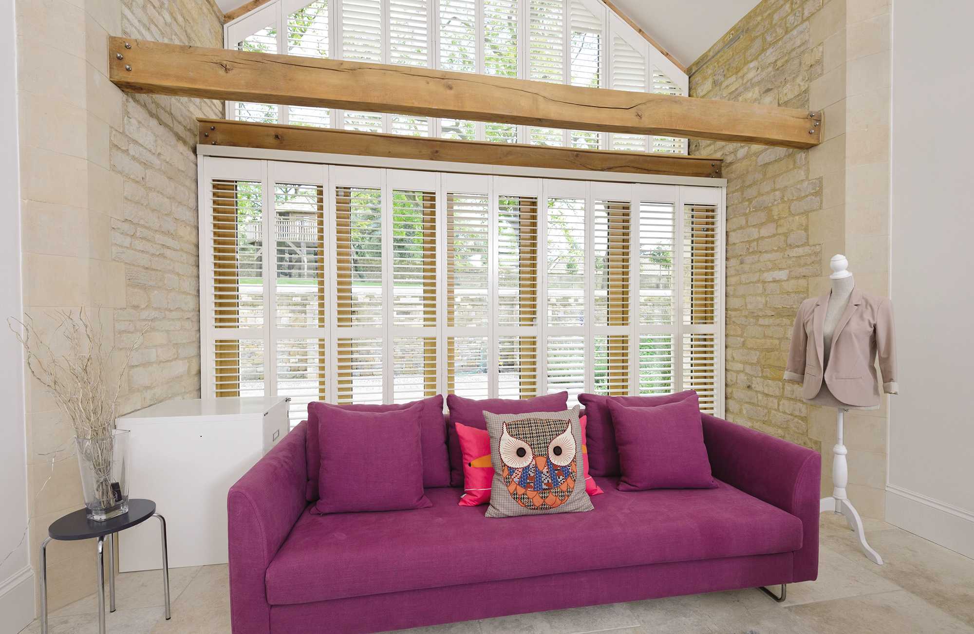 Wide tracked window shutters in white contrasting with a purple sofa in a rustic living room
