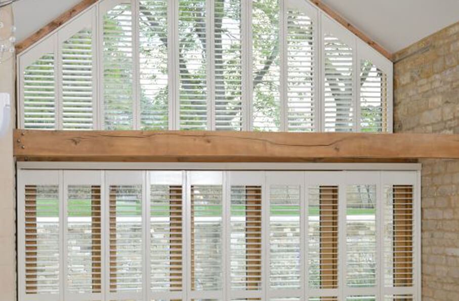 Special shape apex window shutters in white contrasting a brick wall interior with wooden beams.
