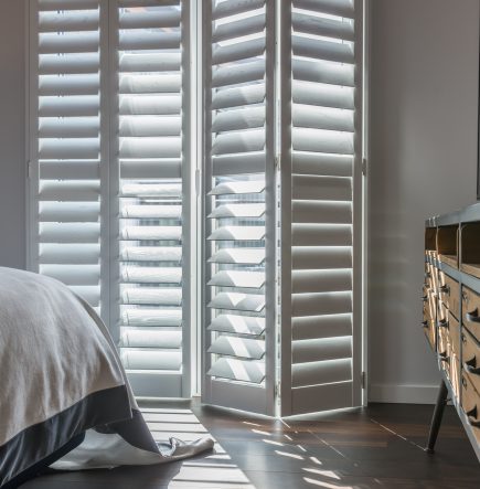 Tracked wooden bedroom window shutters in white complementing a bed and rustic drawers