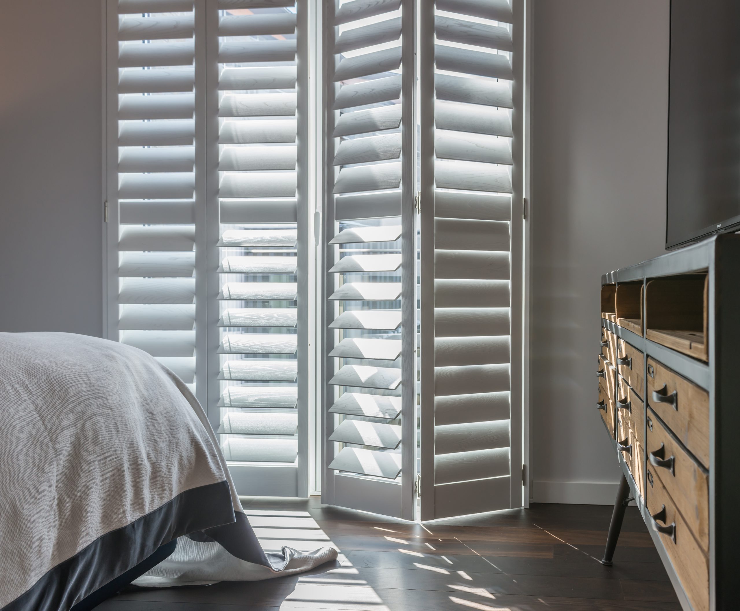 Tracked wooden bedroom window shutters in white complementing a bed and rustic drawers