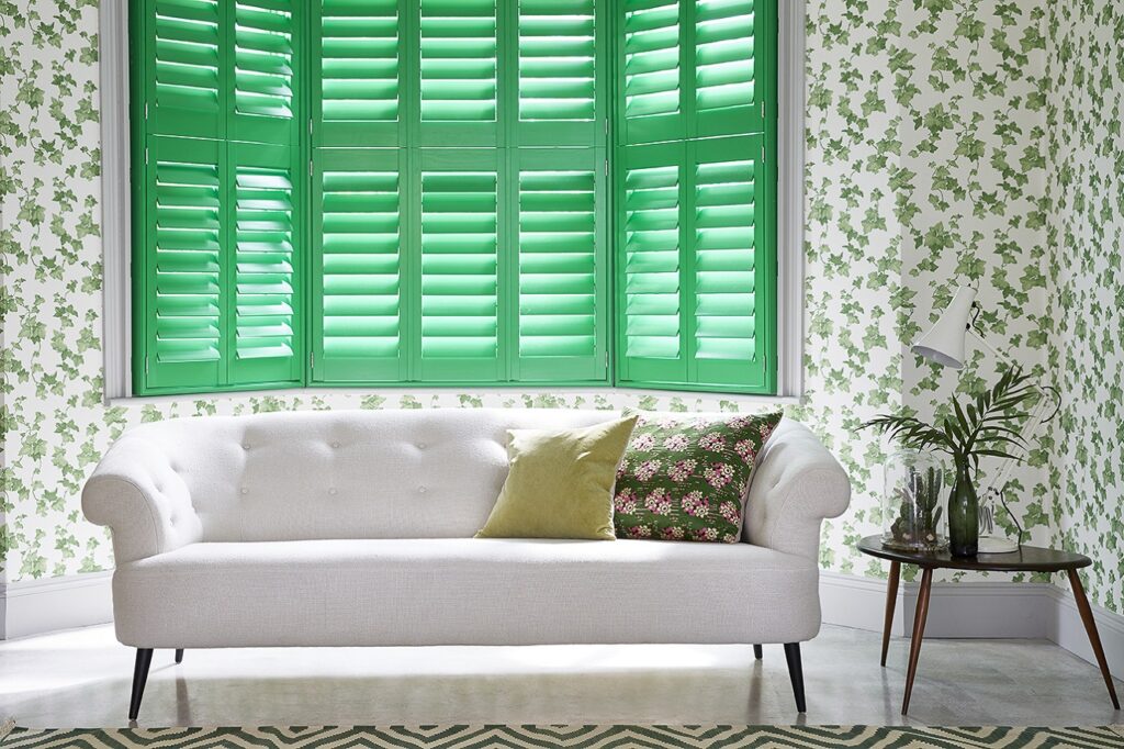 Green bay window shutters in a living room with leafy wallpaper and a white sofa.